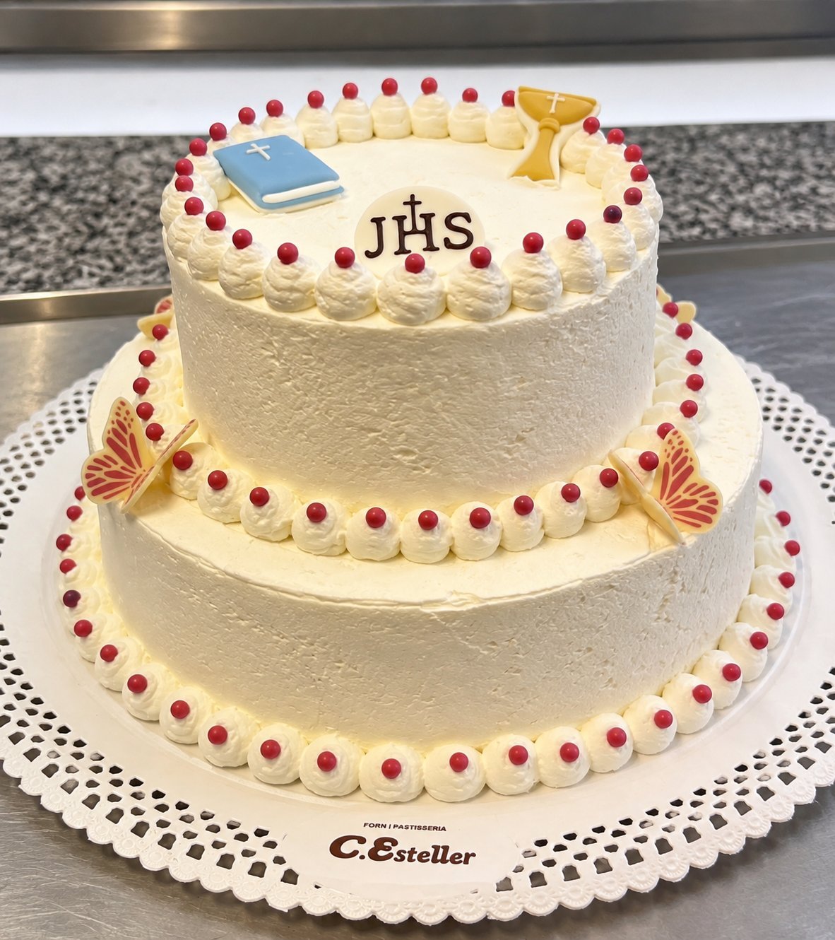 Tarta religiosa de dos pisos con fondant blanco, guirnalda de bayas rojas, mariposas doradas, libro azul, monograma JHS y cáliz dorado, sobre plato con el sello Forn i Pastisseria C. Esteller