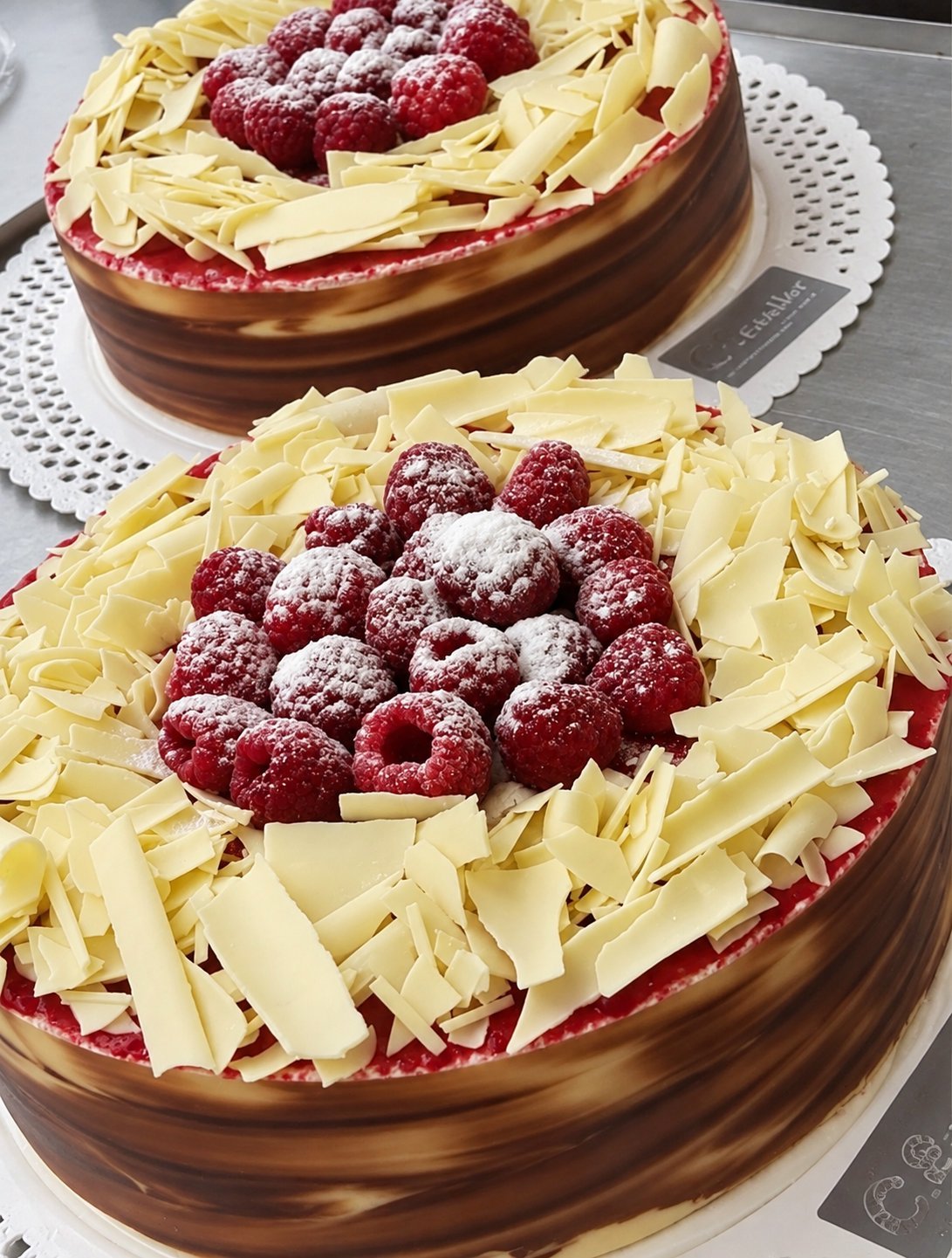 Dos tartas de C. Esteller con capa de chocolate, virutas de chocolate blanco y frambuesas frescas espolvoreadas con azúcar glas sobre plato blanco