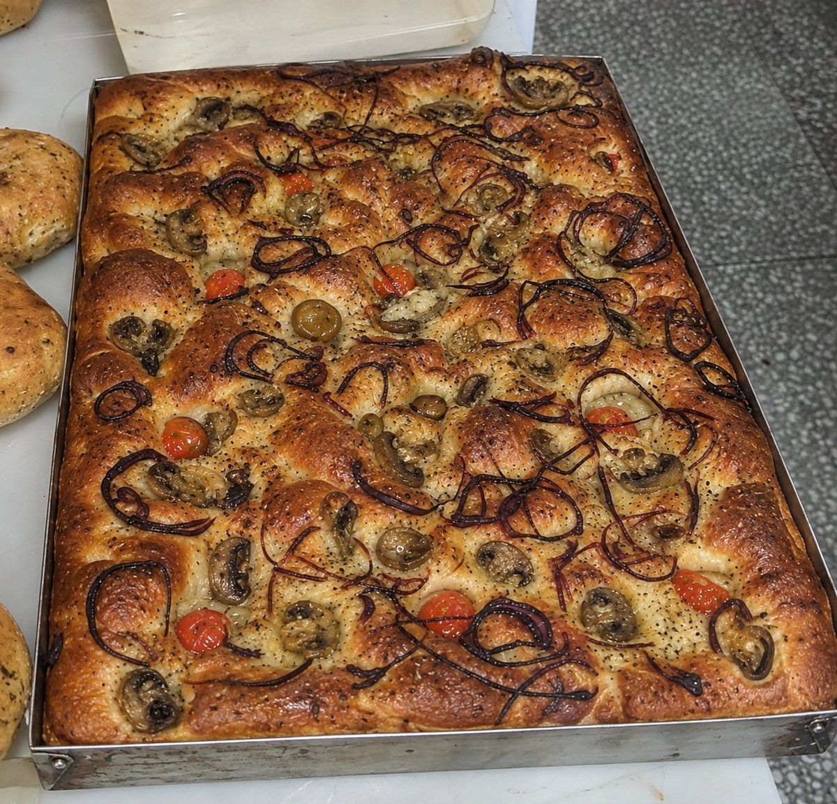 Focaccia recién horneada en bandeja rectangular con cebolla caramelizada, olivas, tomatitos y hierbas aromáticas