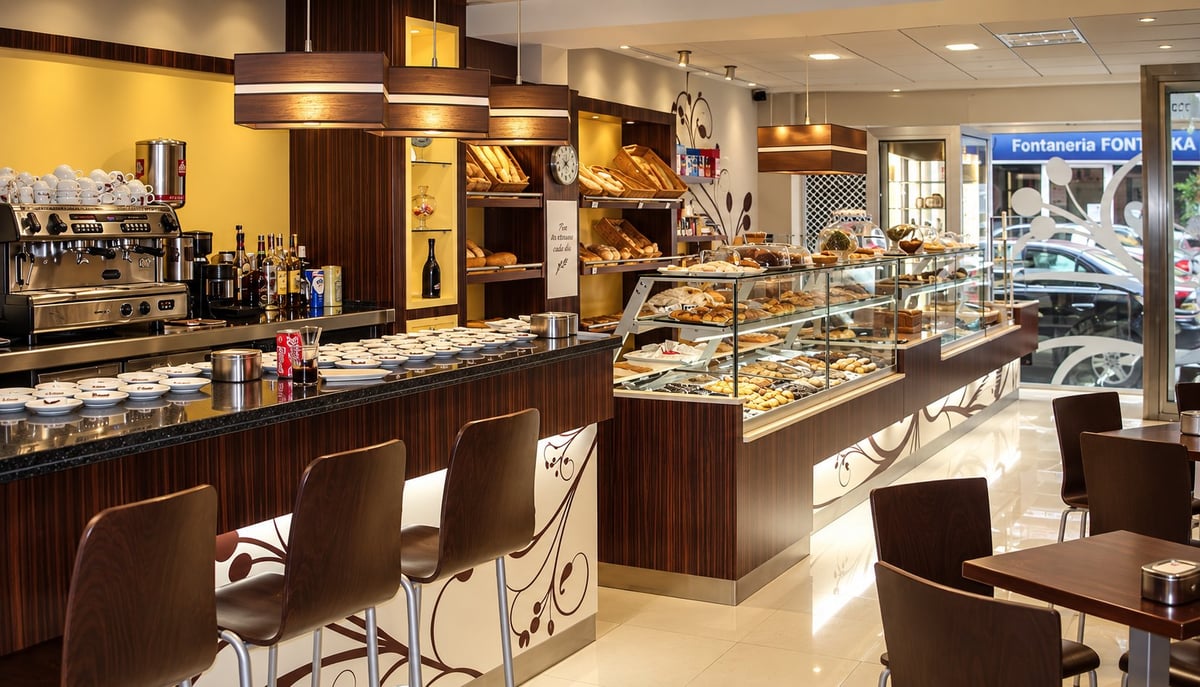 Interior del obrador de C. Esteller con vitrinas de bollería, barra de cafetería con bandejas, sillas de madera y luz cálida en tonos marrón y dorado